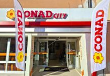 Conad City riapre e aggiorna uno storico presidio di prossimità a Figline Valdarno Entrance of a Conad City supermarket with open glass doors, two tall white flags reading CONAD and a yellow flower logo, and a welcome mat on the floor.