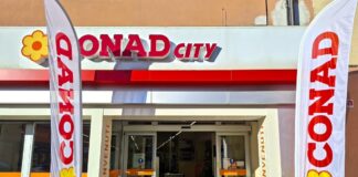 Conad City riapre e aggiorna uno storico presidio di prossimità a Figline Valdarno Entrance of a Conad City supermarket with open glass doors, two tall white flags reading CONAD and a yellow flower logo, and a welcome mat on the floor.