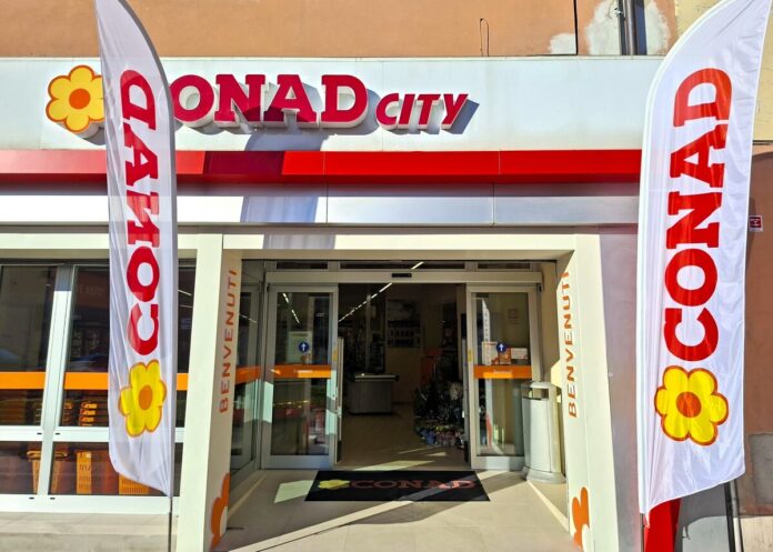 figline cno 1 (1) Entrance of a Conad City supermarket with open glass doors, two tall white flags reading CONAD and a yellow flower logo, and a welcome mat on the floor.